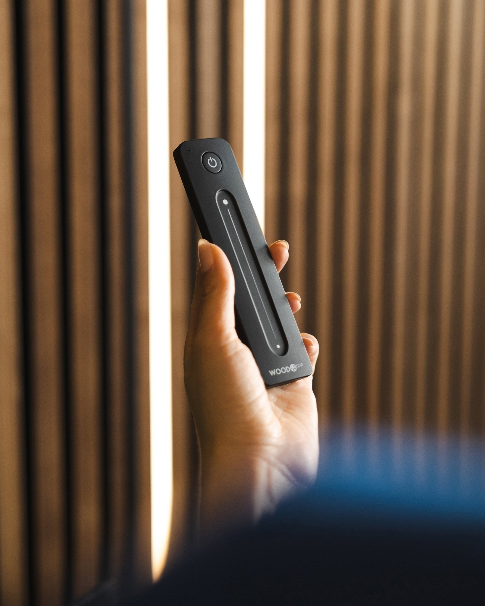 Een hand houdt een black WoodUpp Lumi afstandsbediening voor LED-striplights met aan-uitknop vast, geplaatst voor akoestische lattenwandpanelen van WoodUpp; compatibel met Philips Hue.